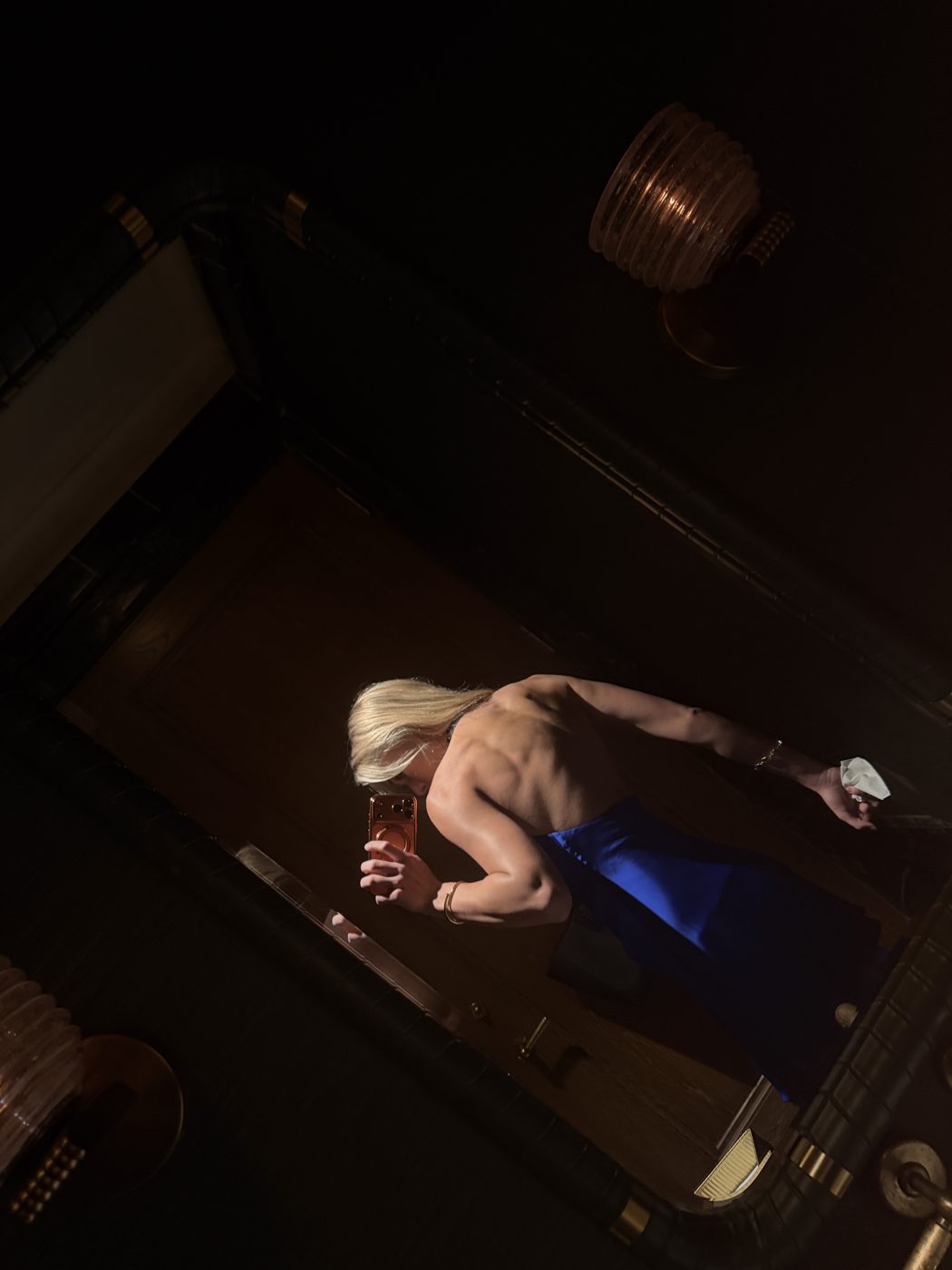 Madeline flexing her back in a dark mirror, in a blue skirt